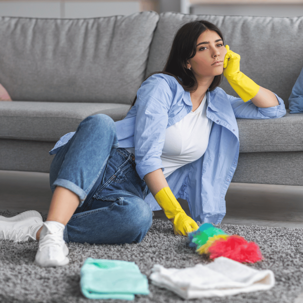 Kvinna sitter på en matta lutandes mot en soffa med flera olika rengöringsmedel och utrustning inför rengöring av plastmatta.