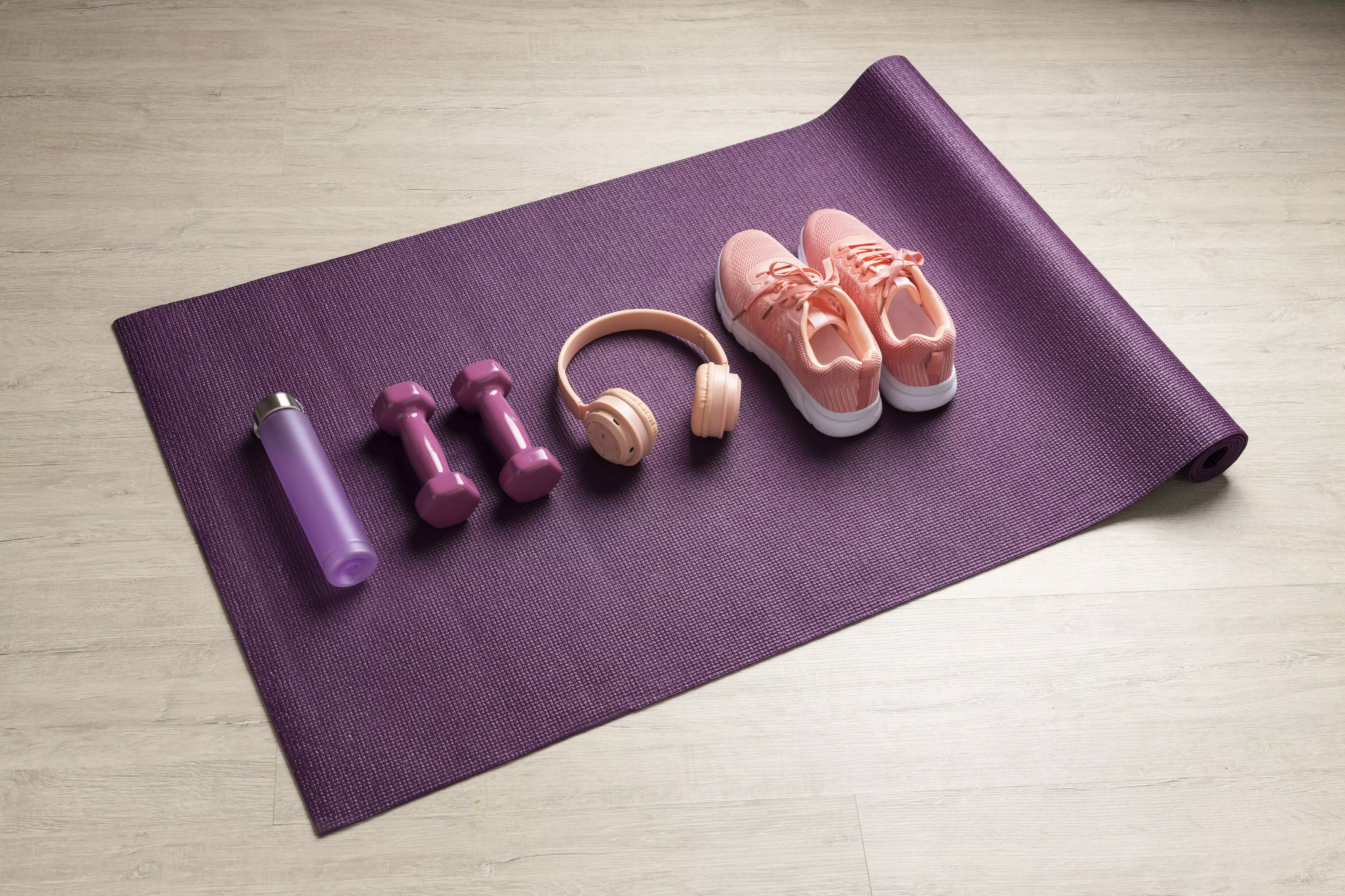 Lila yoga matta för yogaövningar med lila vattenflaska, hantlar, rosa hörlurar och träningsskor.