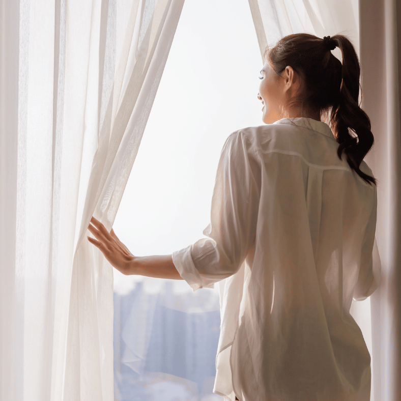 Kvinna tittar ut från fönstret med fina gardiner upphängda.