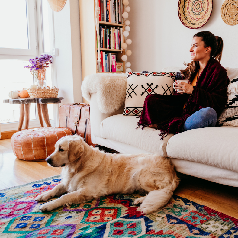 Hund och kvinna ser på TV i vardagsrum. Ljus soffa på en mönstrad matta.