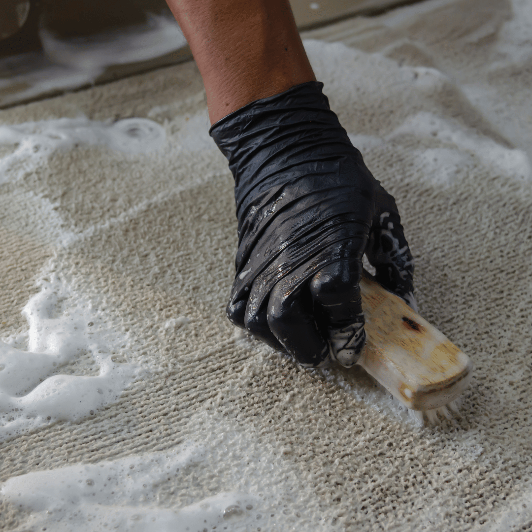 Hand med svart tvätthandske använder en borste och rengöringsmedel för att skrubba smutsig matta.